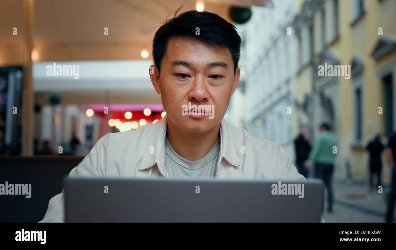 Focalisé asiatique homme touriste assis dehors naviguer sur le réseau social sur ordinateur portable en utilisant le Wi-fi gratuit millénaire homme de voyage blogueur dactylographiant sur l'ordinateur Banque D'Images