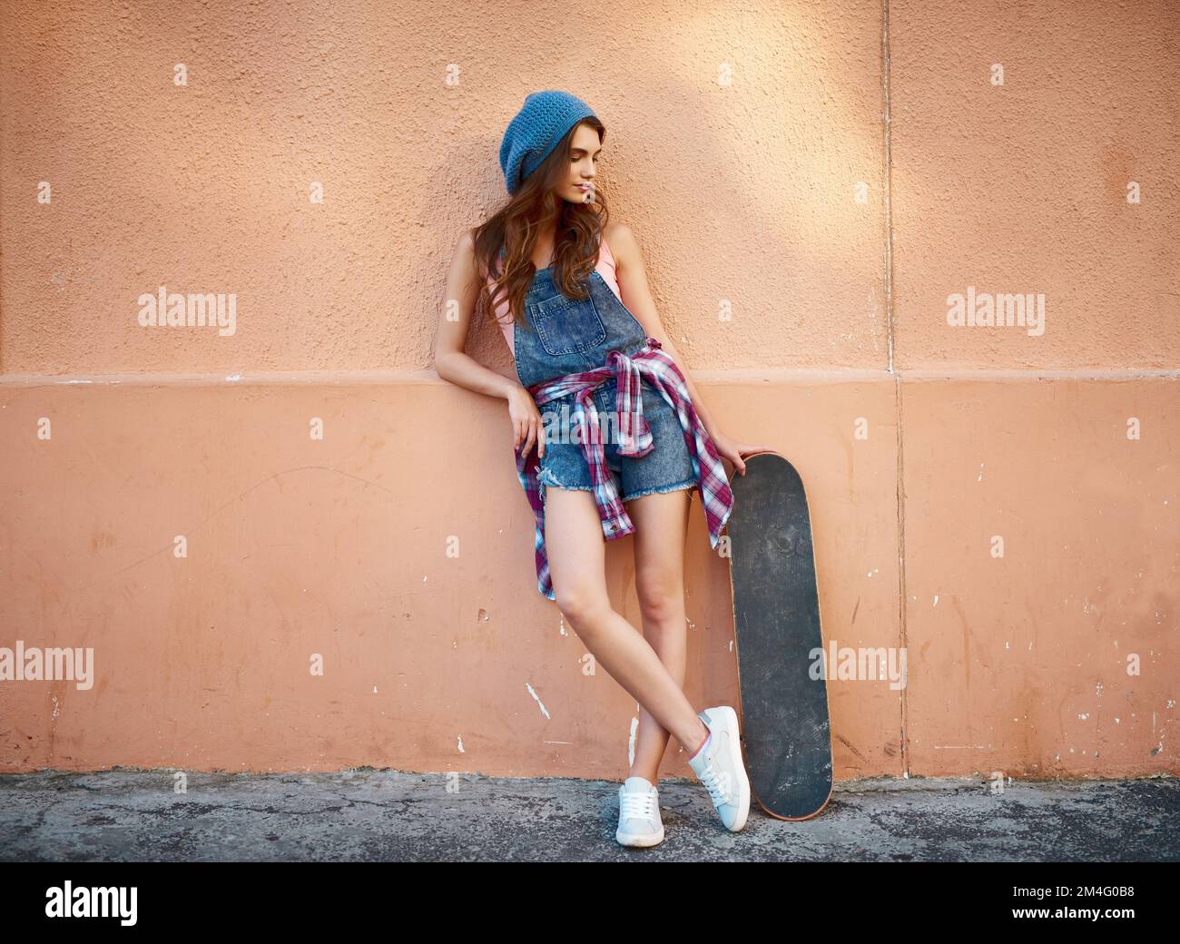 Le skateboarder et sa planche. une jeune femme insouciante debout avec son skateboard sur fond orange. Banque D'Images