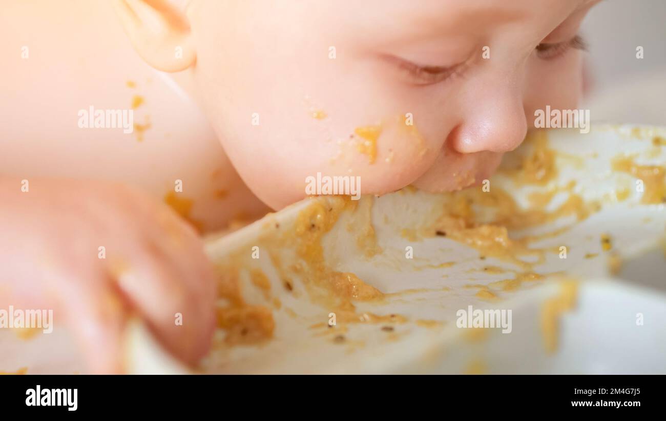 Bébé fille saisit les restes de nourriture de la plaque blanche et mange avec l'expression excitée et préoccupée. L'enfant a le goût de la nourriture sur les mains en se faisant passer le visage et les doigts Banque D'Images