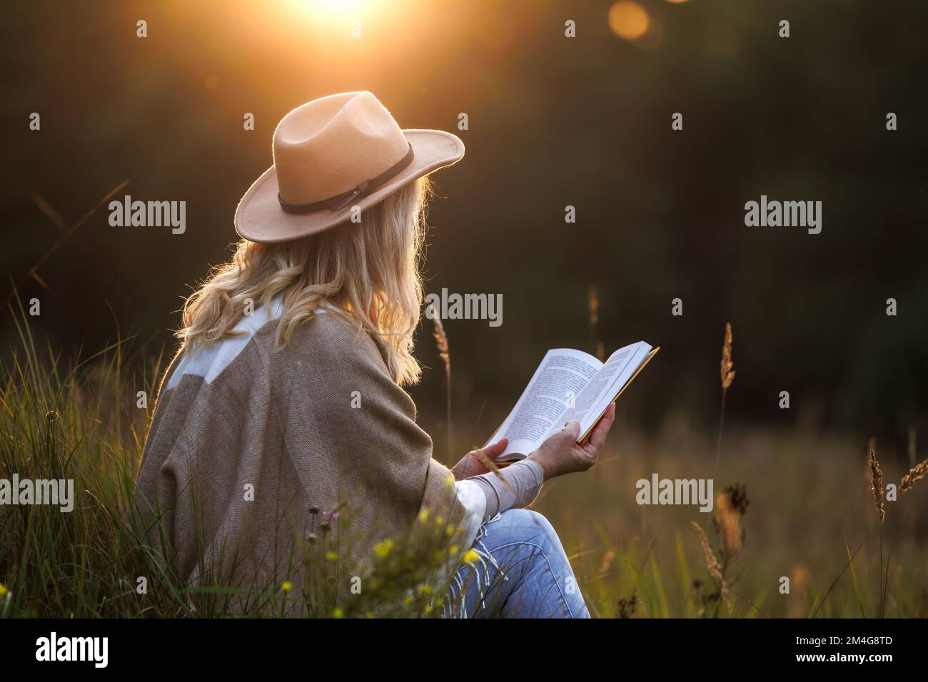 Femme avec chapeau et poncho lisant le livre à l'extérieur au coucher du soleil. Détente et détox numérique dans la nature Banque D'Images