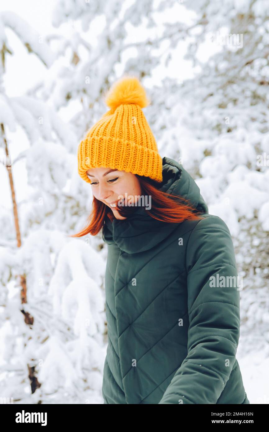 Fille gaie avec des cheveux rouges dans des vêtements chauds jouant avec la neige à l'extérieur près de la belle forêt Banque D'Images