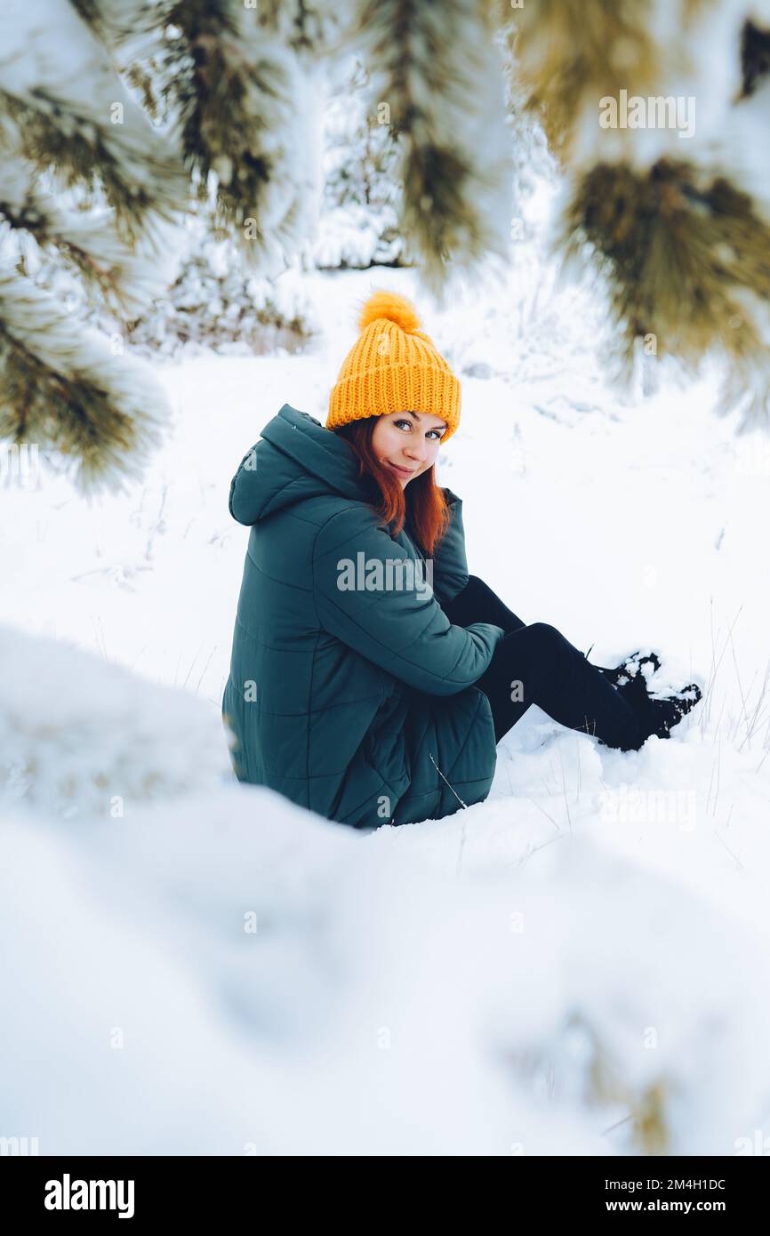 Fille gaie avec des cheveux rouges dans des vêtements chauds jouant avec la neige à l'extérieur près de la belle forêt Banque D'Images