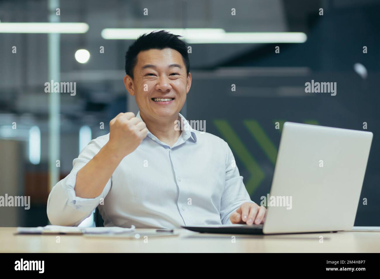 Un étudiant asiatique assis au bureau avec un ordinateur portable, a obtenu de bonnes nouvelles, lettre en ligne de l'université. passé l'examen, entrée à l'université. Il regarde joyeusement la caméra, montre un geste victorieux. Banque D'Images