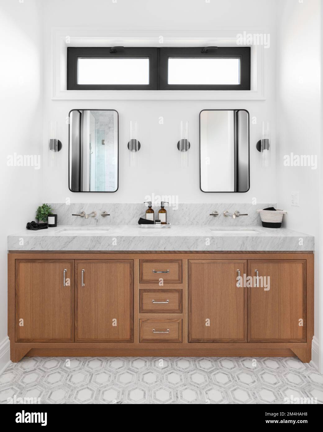 Une salle de bains confortable avec un meuble en bois, un comptoir en marbre, des sconces autour de miroirs carrés et un beau sol carrelé. Banque D'Images