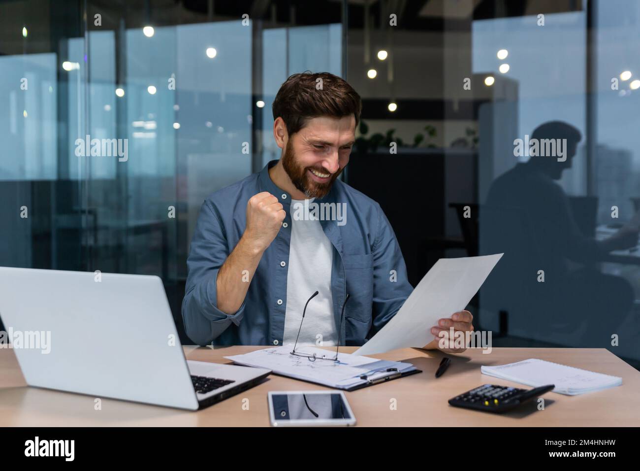 Homme d'affaires réussi dans une chemise décontractée faisant de la paperasse, patron avec la barbe et les lunettes assis au bureau sur le lieu de travail en utilisant l'ordinateur portable travaillant avec des documents, tenant main vers le haut célébrant la victoire et le triomphe. Banque D'Images