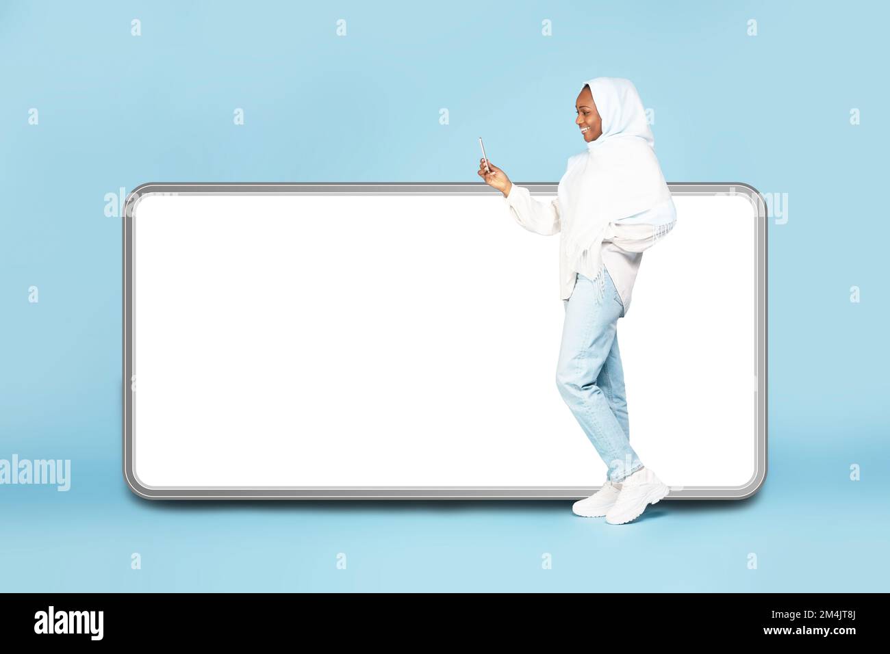 Femme noire excitée dans hijab debout près de grand téléphone portable avec écran vide et en utilisant un appareil mobile, maquette pour la publicité Banque D'Images