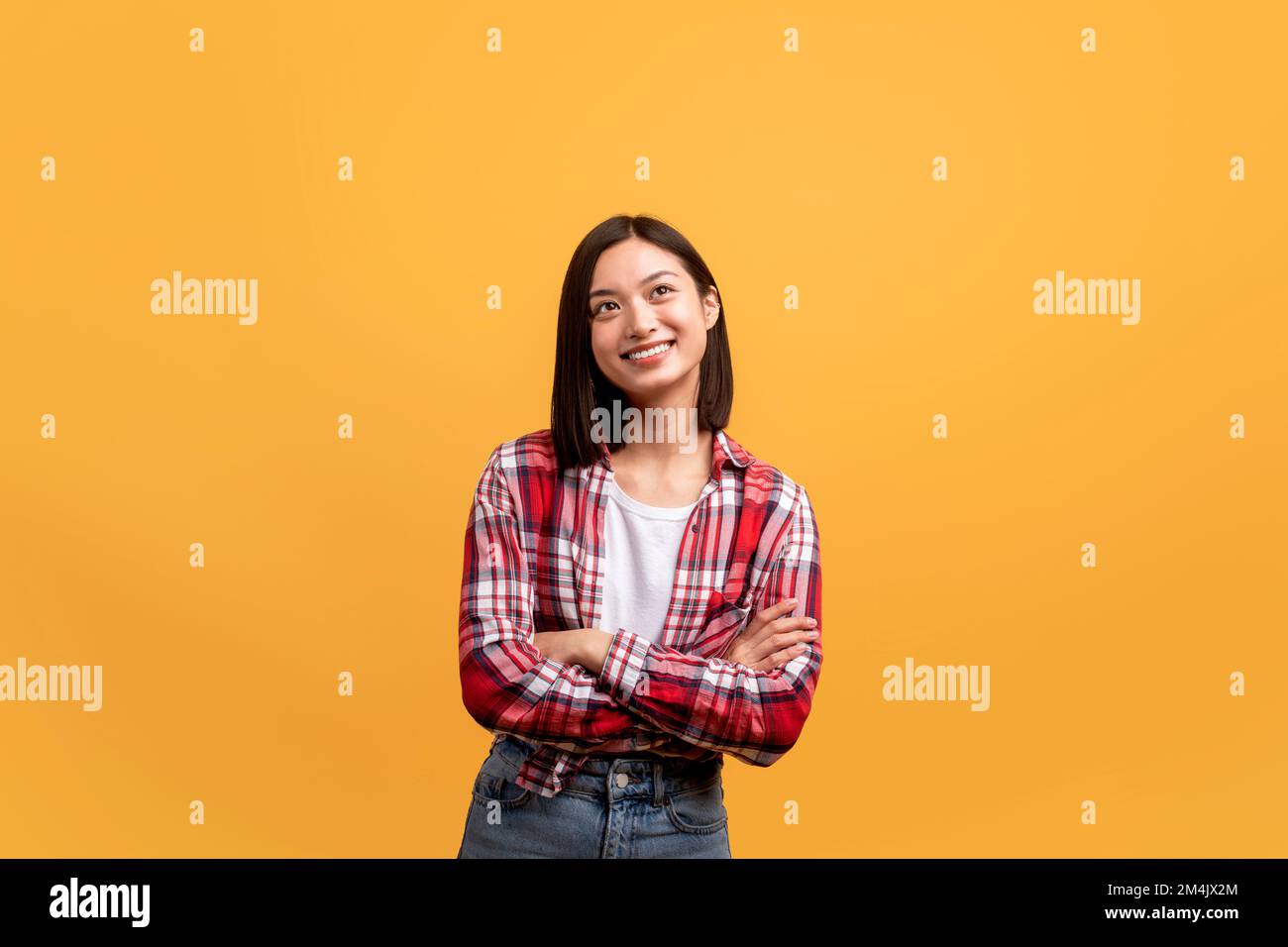 Femme asiatique positive rêveuse debout avec les bras pliés et regardant de côté, posant sur fond jaune, espace de copie Banque D'Images