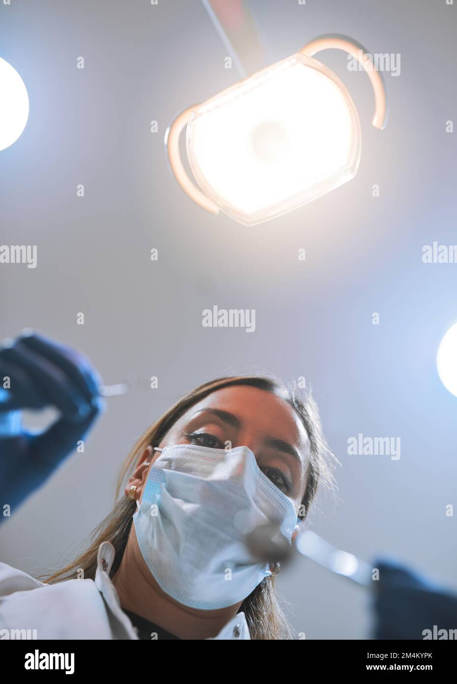 Ouverture large. Plan pov à angle bas d'une jeune femme dentiste focalisée portant un masque chirurgical tout en essayant de travailler sur les dents d'un patient. Banque D'Images