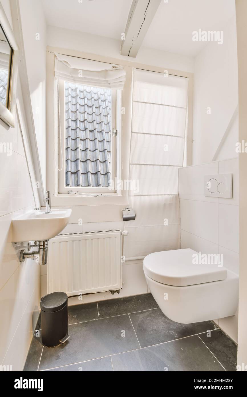 une salle de bains avec toilettes, lavabo et fenêtre dans le coin à côté est une poubelle noire sur le sol Banque D'Images