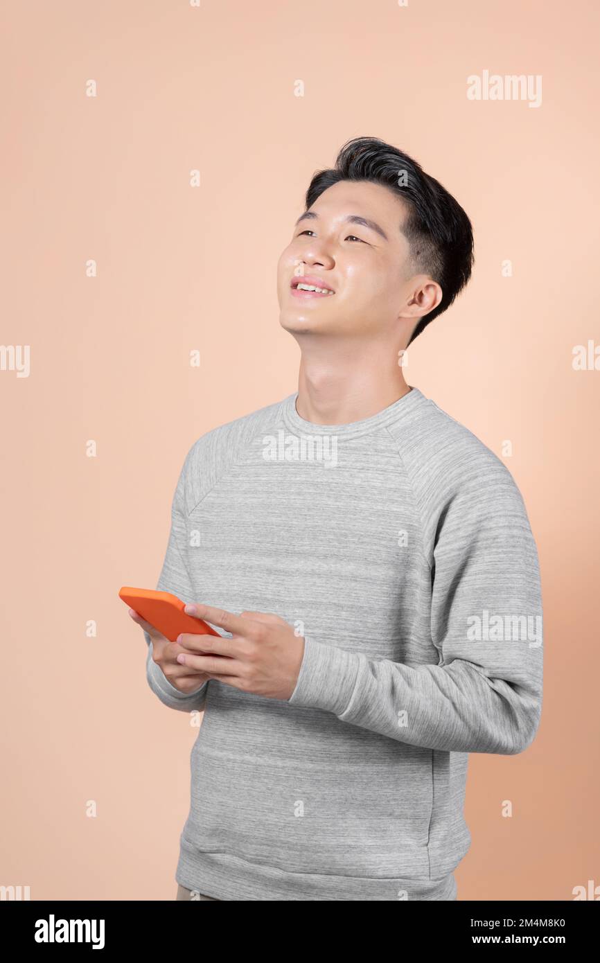 Jeune homme asiatique utilisant un téléphone portable isolé sur fond beige regardant vers le haut tout en souriant Banque D'Images