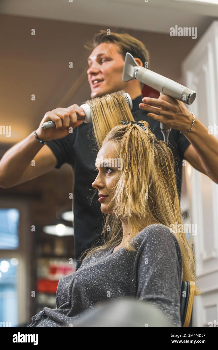 Rendez-vous avec un coiffeur. Jeune femme blonde ayant ses cheveux soufflés séchés par un coiffeur professionnel. Mise au point au premier plan. Portrait vertical en intérieur. Copier l'espace. Photo de haute qualité Banque D'Images