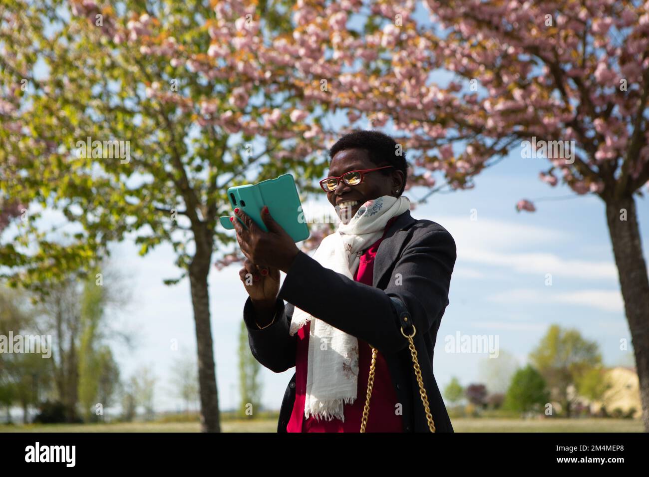 Élégante femme africaine souriante utilisant un smartphone dans un parc extérieur. Nature et arbres fleuris le jour ensoleillé du printemps. Concept d'utilisation de la technologie dans le Banque D'Images