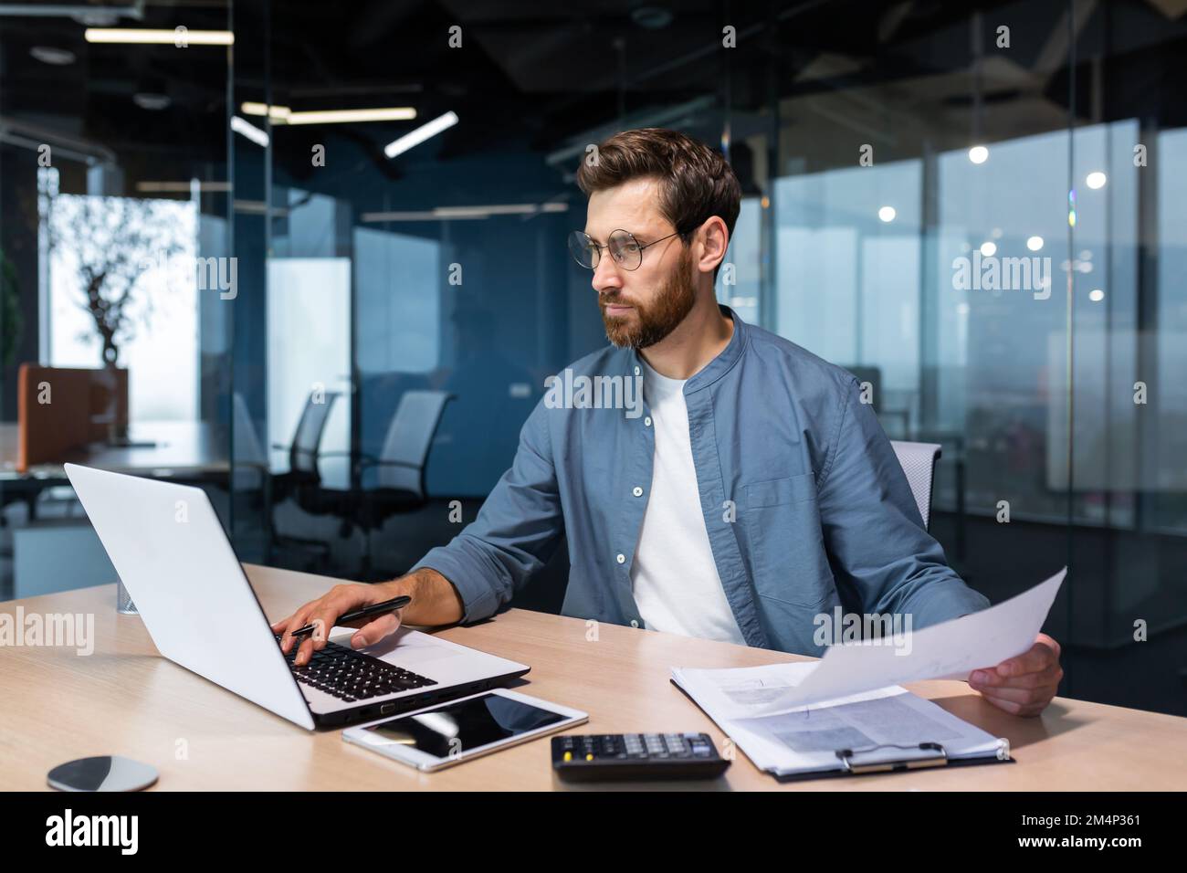 Un sérieux jeune homme comptable, financier, analyste, auditeur est assis dans le bureau à la table. Il tient des documents et un stylo entre ses mains, vérifie des comptes, des finances, des types sur un ordinateur portable. Banque D'Images