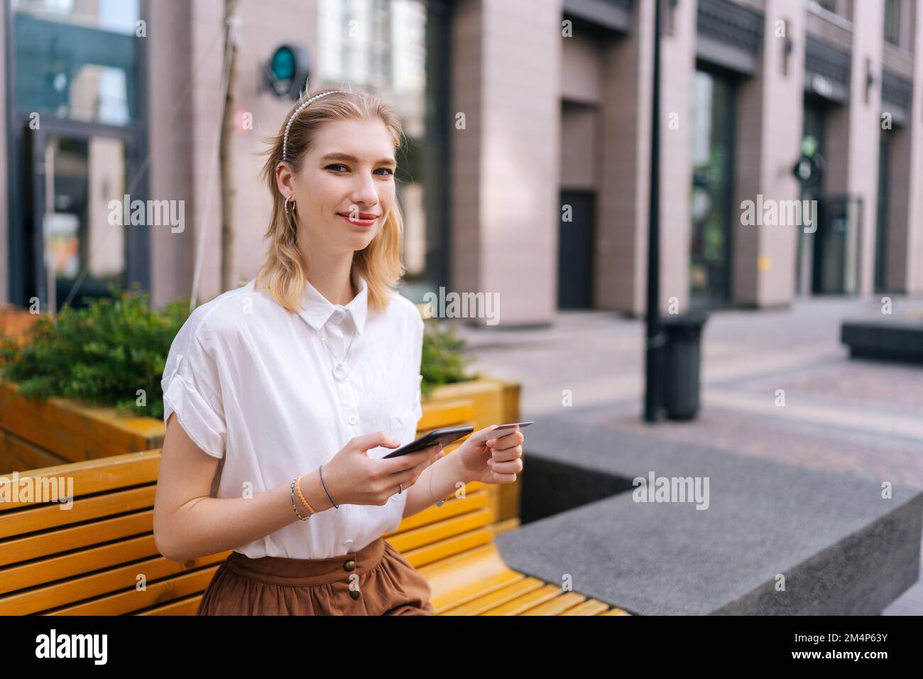Prise de vue moyenne d'une jeune femme positive tenant une carte de crédit et mobile dans les mains souriant regardant la caméra assis sur un banc sur la rue de la ville. Banque D'Images
