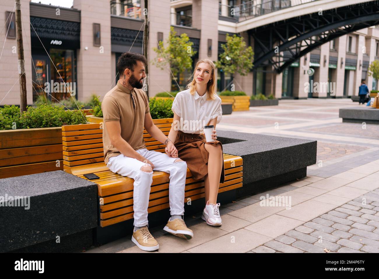 Photo d'un jeune couple aimant assis sur un banc, tenant les mains et prenant un café à emporter en profitant du temps passé ensemble à l'extérieur. Banque D'Images