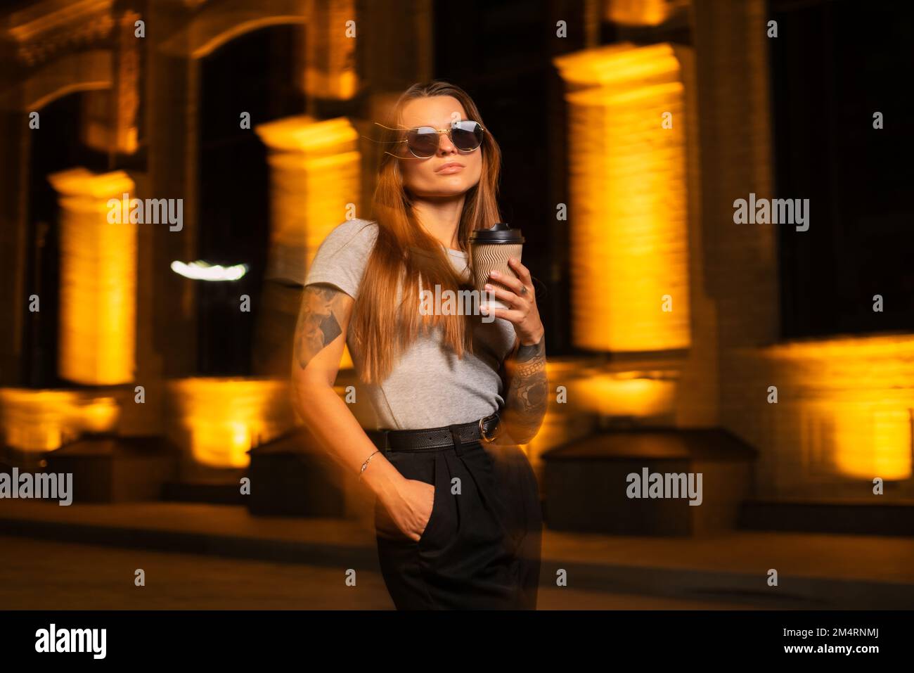 Portrait dans le mouvement flou d'une jeune femme moderne et attrayante portant des lunettes et tenant une tasse de café la nuit dehors Banque D'Images