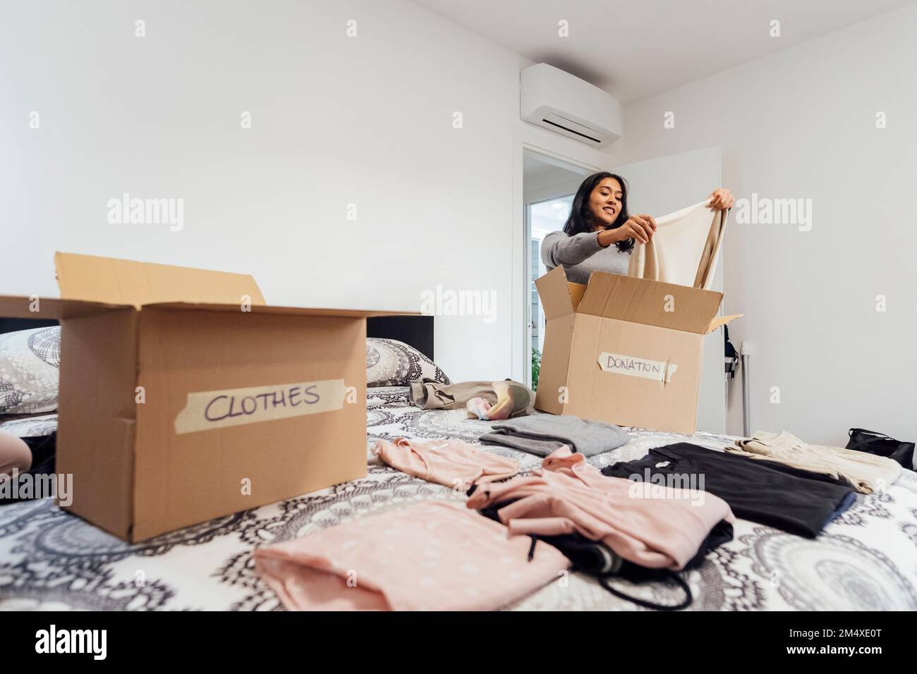 Jeune femme souriante emballant une boîte de vêtements à donner sur le lit Banque D'Images