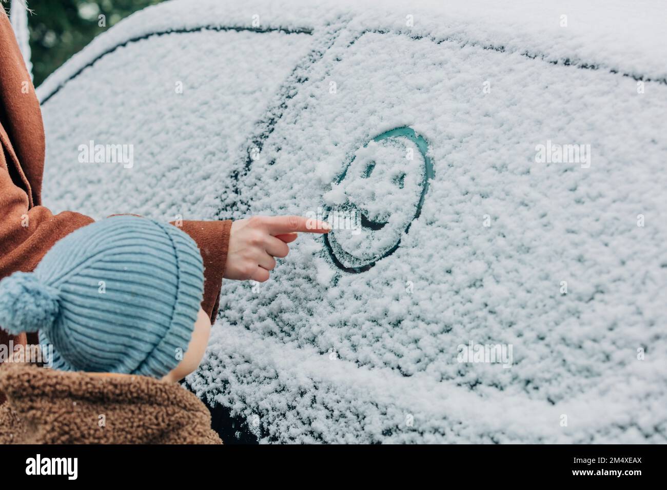 Garçon debout par la mère dessinant le visage humain sur la fenêtre de voiture recouverte de neige Banque D'Images
