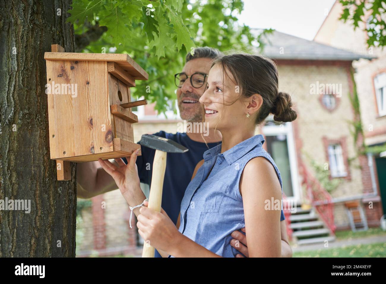 Fille souriante avec un père suspendu boîte de nid sur le tronc de l'arbre dans la cour arrière Banque D'Images