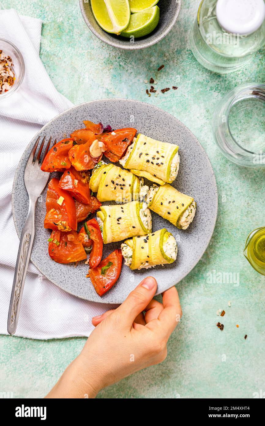 Point de vue personnel de la femme ramassant une assiette de courgettes et de rolls de ricotta avec une salade de tomates Banque D'Images