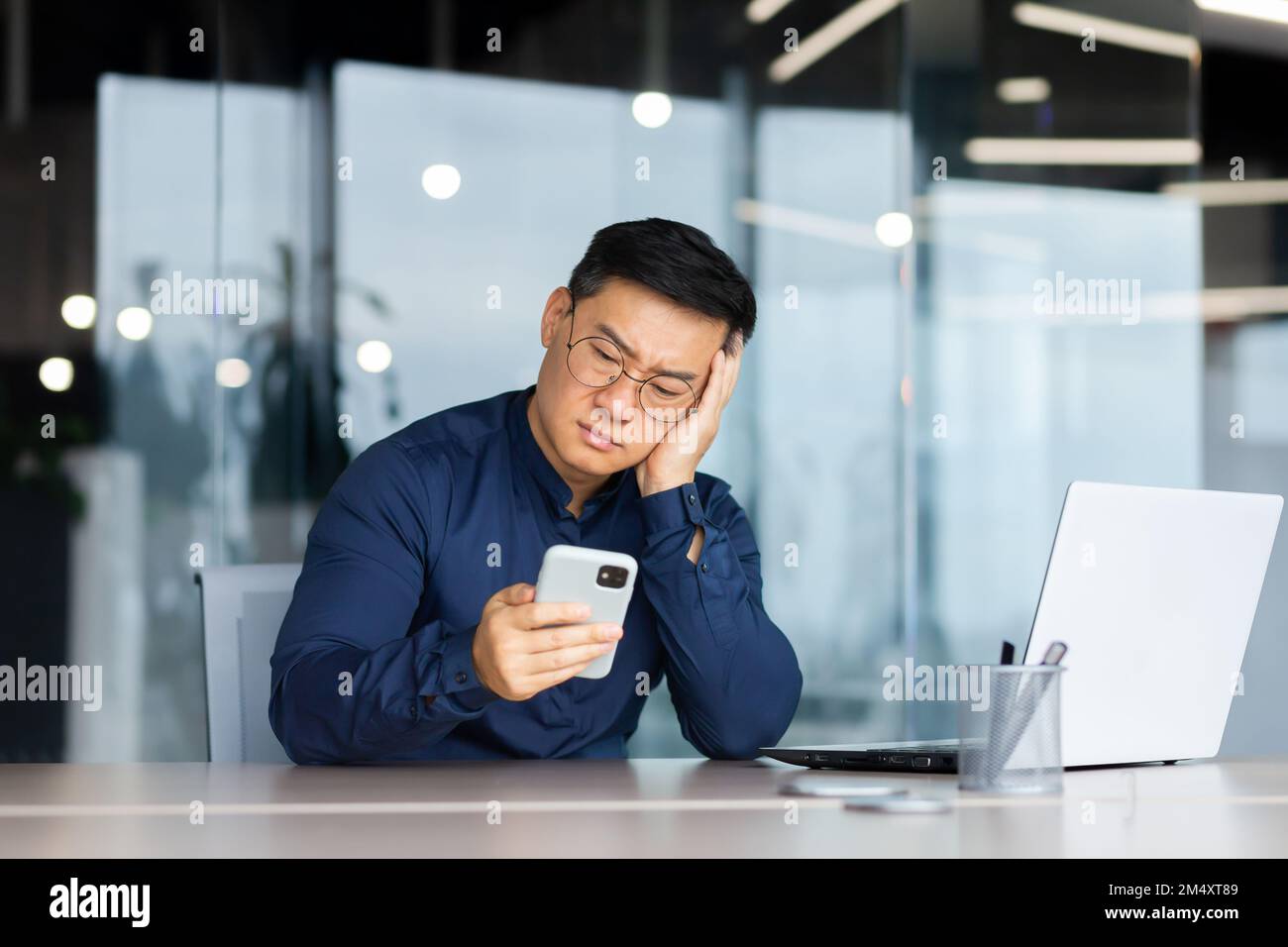 Portrait d'un jeune asiatique inquiet assis dans le bureau à la table, tenant sa tête avec sa main, regardant malheureusement le téléphone. S'ennuyer, a reçu de mauvaises nouvelles, des messages. Banque D'Images