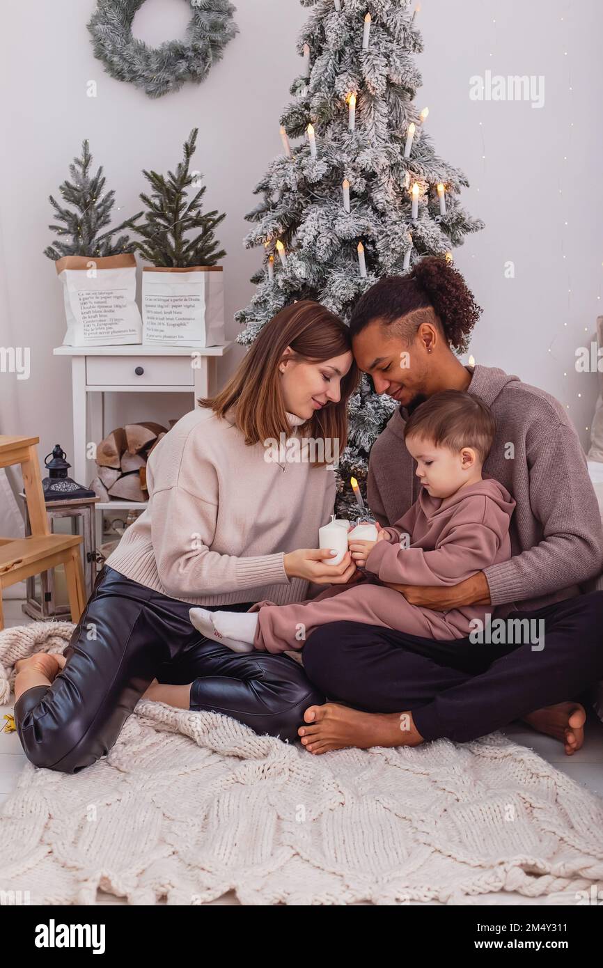 Une jeune famille multiraciale avec un enfant est assise sur une couverture tricotée près d'un arbre de Noël Banque D'Images