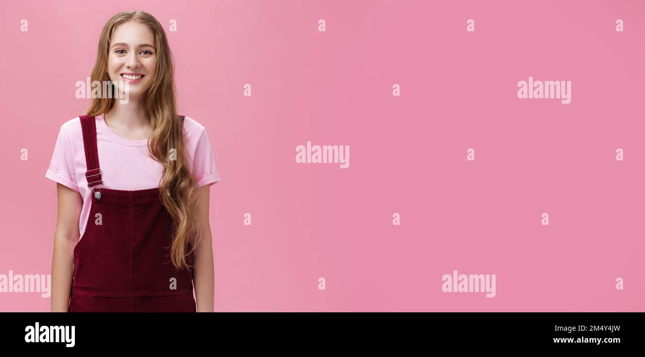 Portrait de charmante petite fille mince avec de longs cheveux justes dans des combinaisons en velours côtelé souriant debout joyeusement debout avec sourire positif de sourire posé contre le rose Banque D'Images