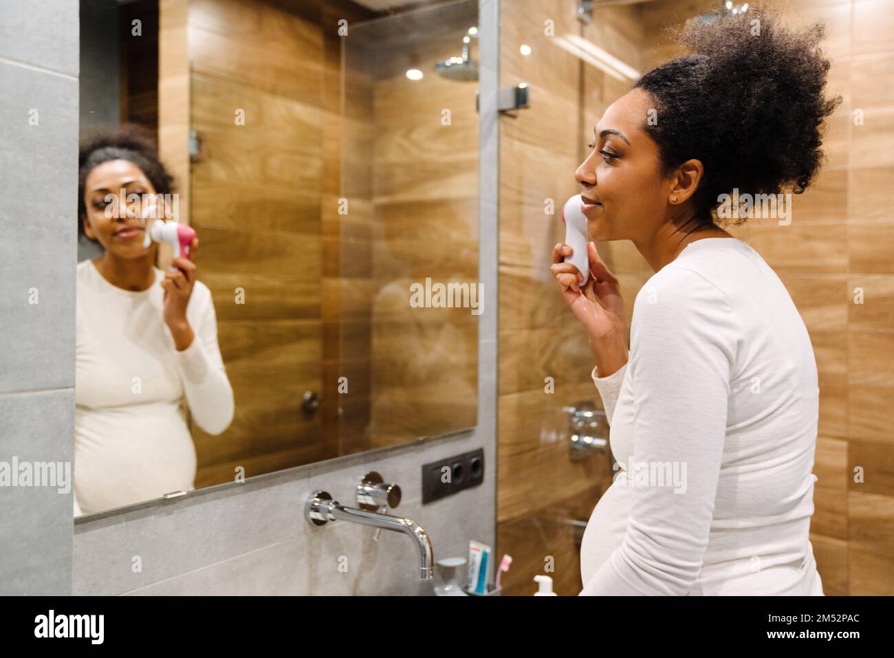 Concept beauté et soin de la peau. Jeune femme africaine utilisant de la mousse nettoyante pour laver son visage avec une brosse en silicone devant le miroir dans la salle de bains Banque D'Images