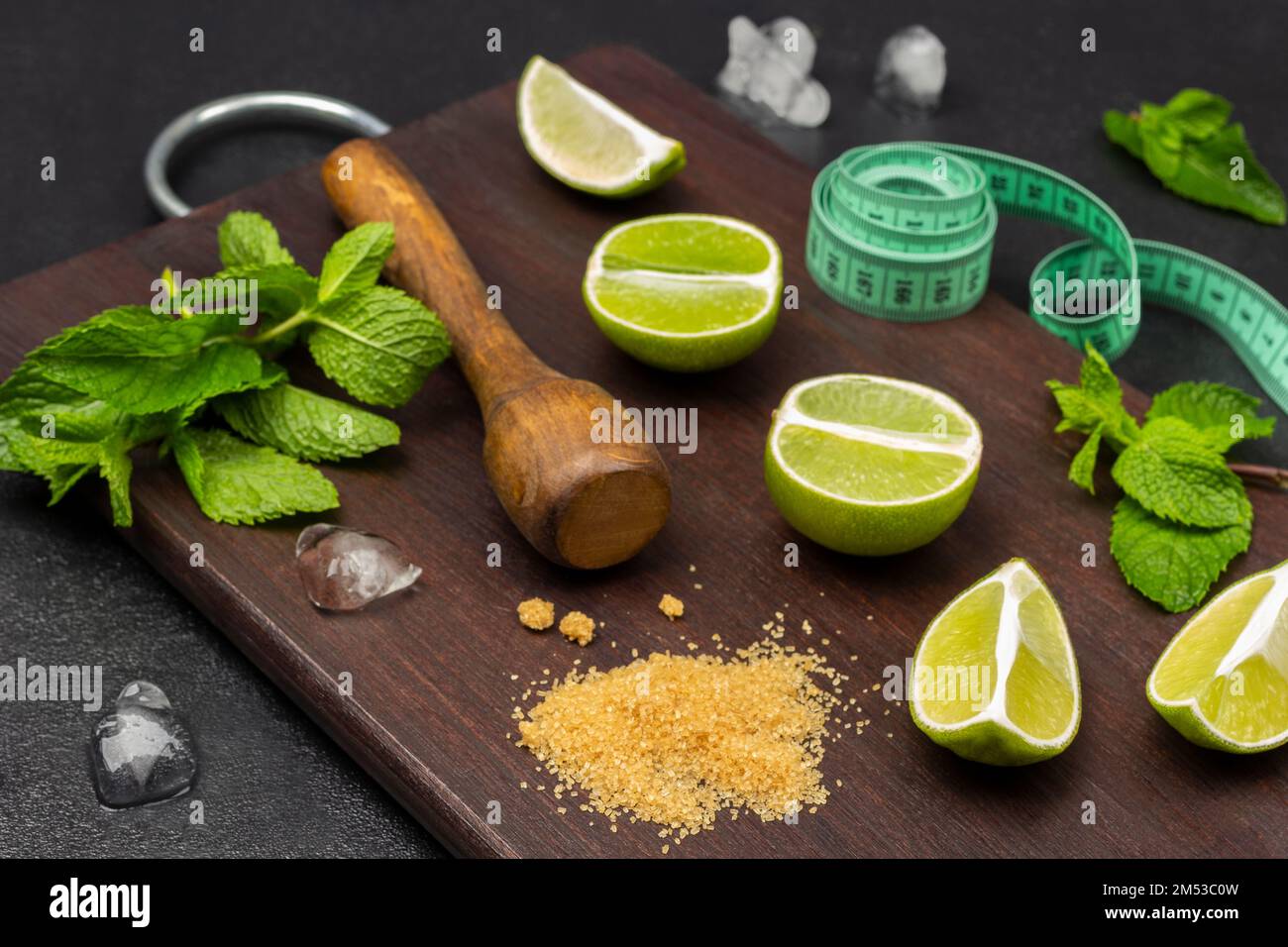 Demi-morceaux de lime, branches de menthe, pilon en bois et ruban de mesure sur une planche. Morceaux de glace sur la table. Vue de dessus. Arrière-plan en bois sombre. Banque D'Images