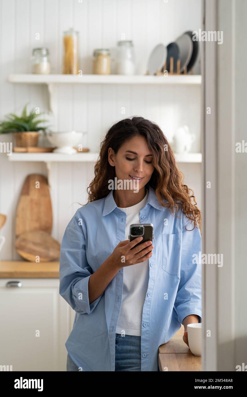 Jeune femme souriante agréable debout dans la cuisine tenant un smartphone, regardant la cuisine vidéo recette Banque D'Images
