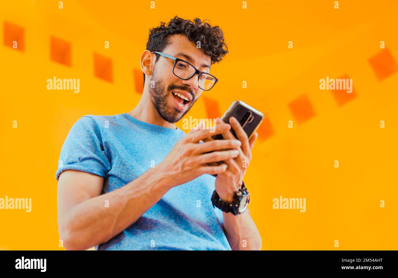 Joyeux gars envoyant un message avec un téléphone cellulaire près d'un mur jaune à l'extérieur. Style de vie de beau barbu homme utilisant le téléphone cellulaire à l'extérieur, angle bas Banque D'Images