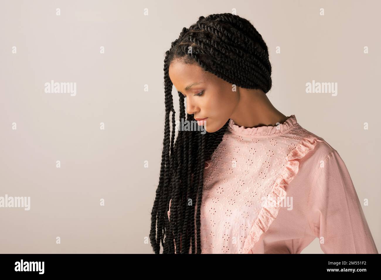 Jeune femme noire en blouse rétro rose avec de longues tresses afro regardant vers le bas sur fond gris Banque D'Images