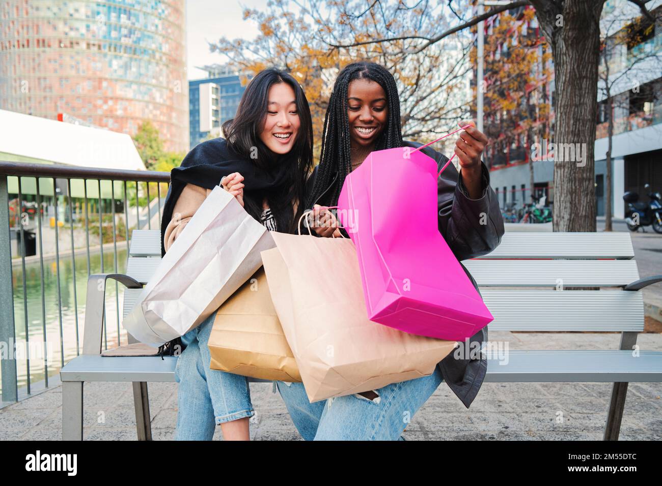 Deux jeunes femmes multiraciales souriant et regardant à l'intérieur d'un sac de shopping assis sur un banc. Quelques amis heureux montrant leurs achats sur une semaine de vente. Concept de consommables. Photo de haute qualité Banque D'Images