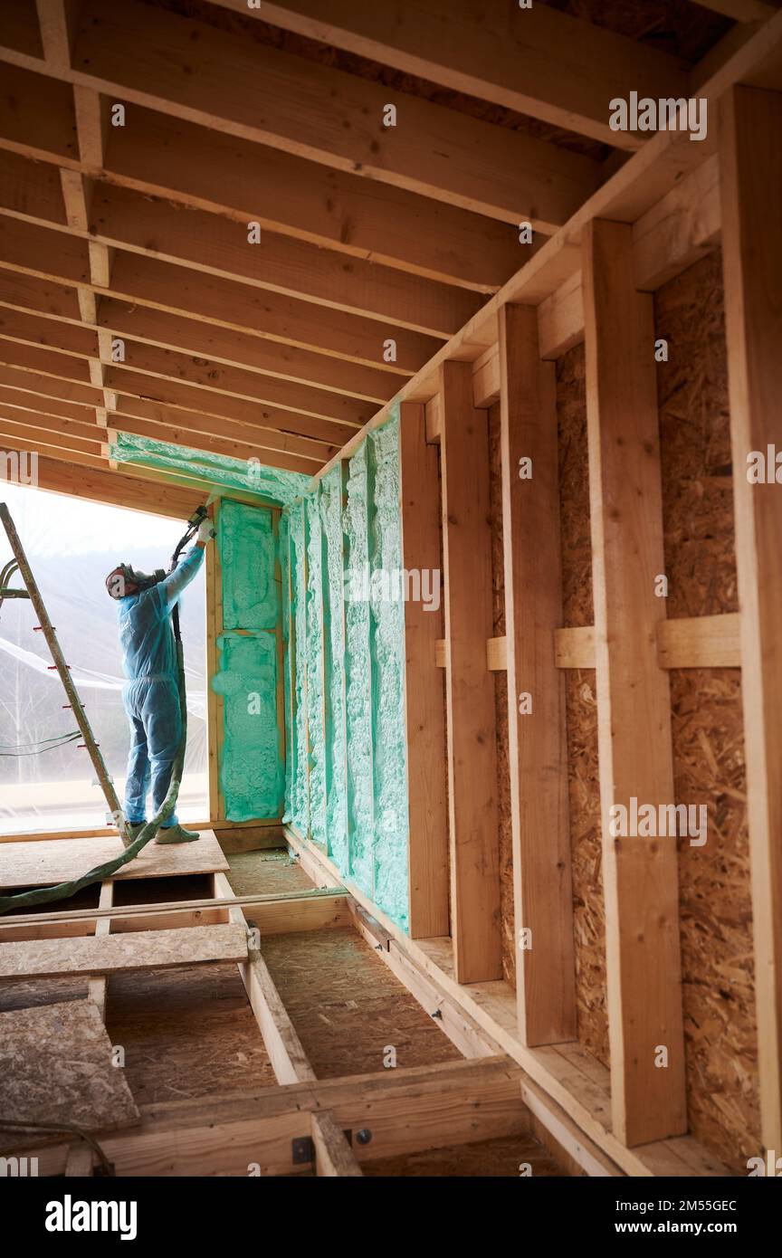 Maison en bois isolante de construction mâle. Homme ouvrier pulvérisant de la mousse de polyuréthane à l'intérieur de future maison, à l'aide d'un pistolet à composant plural. Concept de construction et d'isolation. Banque D'Images