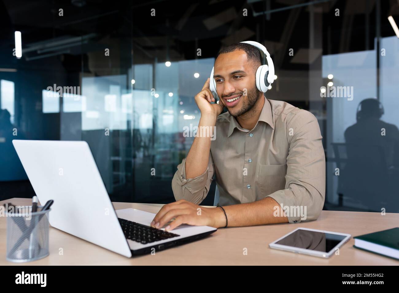 Jeune homme d'affaires hispanique en chemise travaillant au bureau avec un ordinateur portable au travail, homme avec un casque à l'écoute de musique et de podcasts audio au travail. Banque D'Images