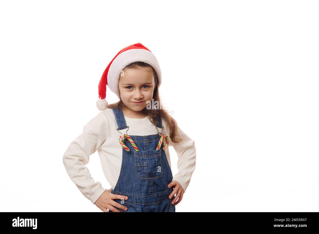 Adorable enfant fille portant un chapeau de père Noël, posant avec des cannes de bonbons sur la poche de ses salopettes en denim, fond blanc Banque D'Images