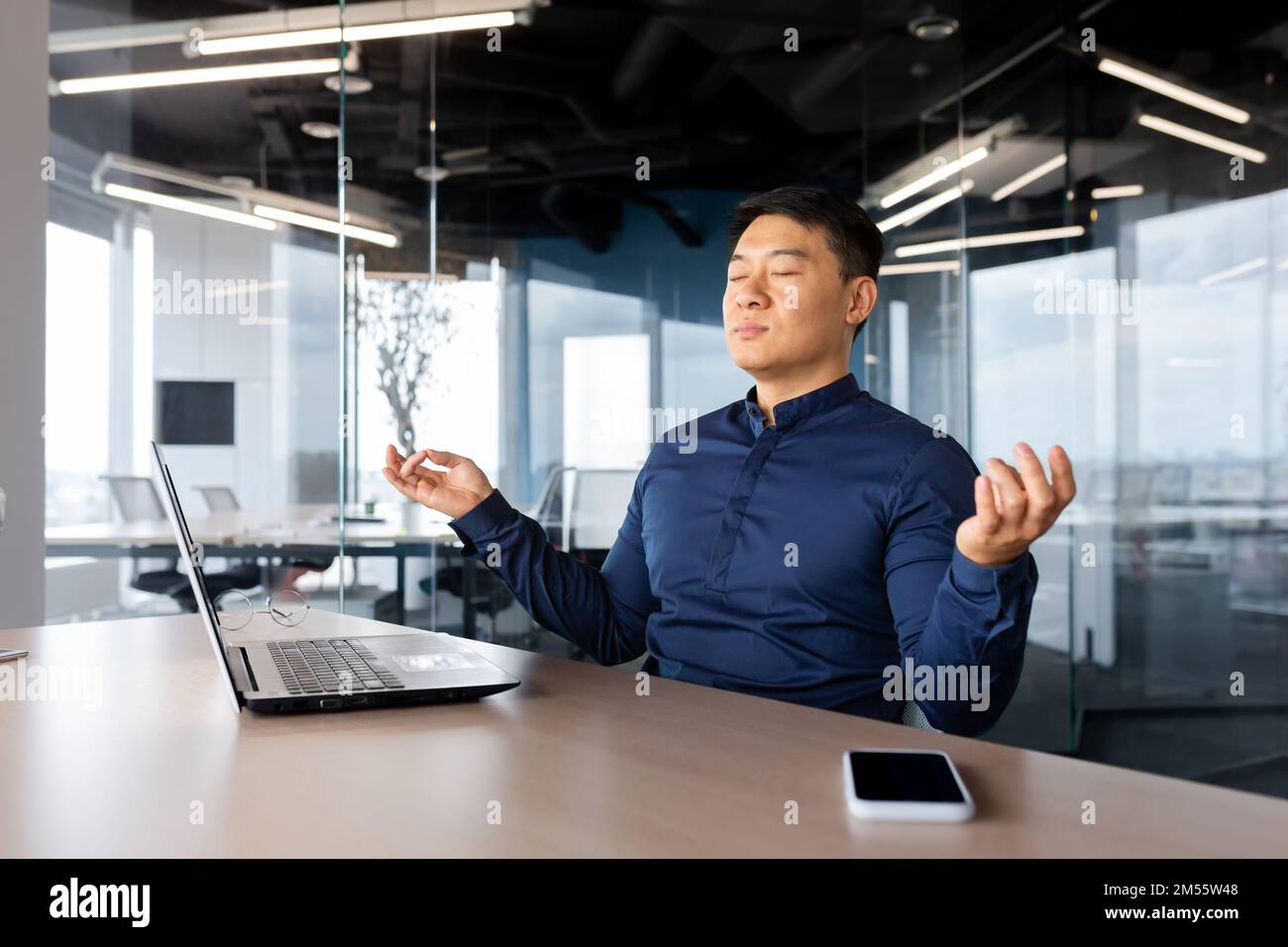 Jeune homme d'affaires asiatique fatigué méditant au travail. Il s'assoit à la table du bureau, prend une pause, ferme les yeux, les mains en position lotus, se détend. Banque D'Images