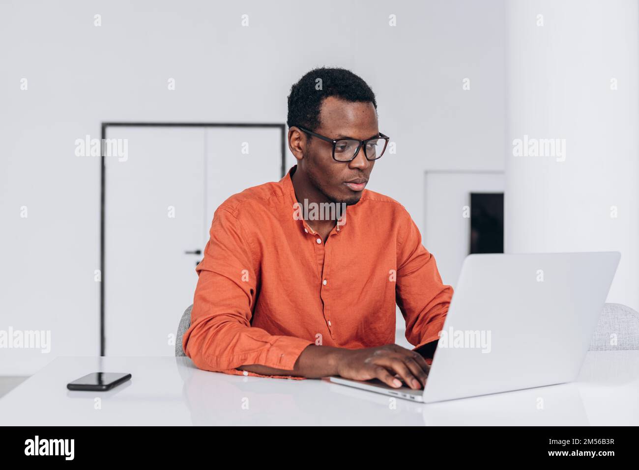 Un jeune homme afro-américain concentré en vêtements et lunettes orange travaille sur un ordinateur portable moderne dans un espace de travail confortable Banque D'Images