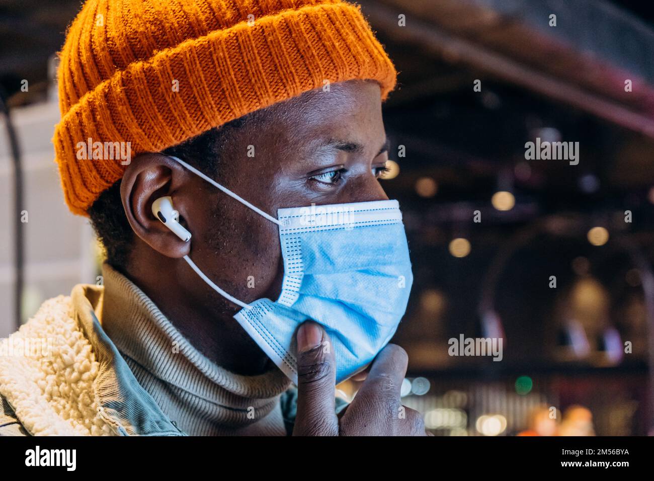 Joli homme afro-américain en chapeau avec masque de protection et écouteurs sans fil touche le menton sur un arrière-plan flou gros plan Banque D'Images