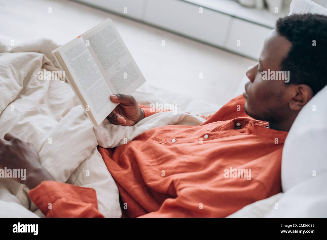 Beau jeune homme noir en pyjama lit un livre intéressant allongé dans un lit confortable avec oreillers doux et une couverture dans la chambre Banque D'Images