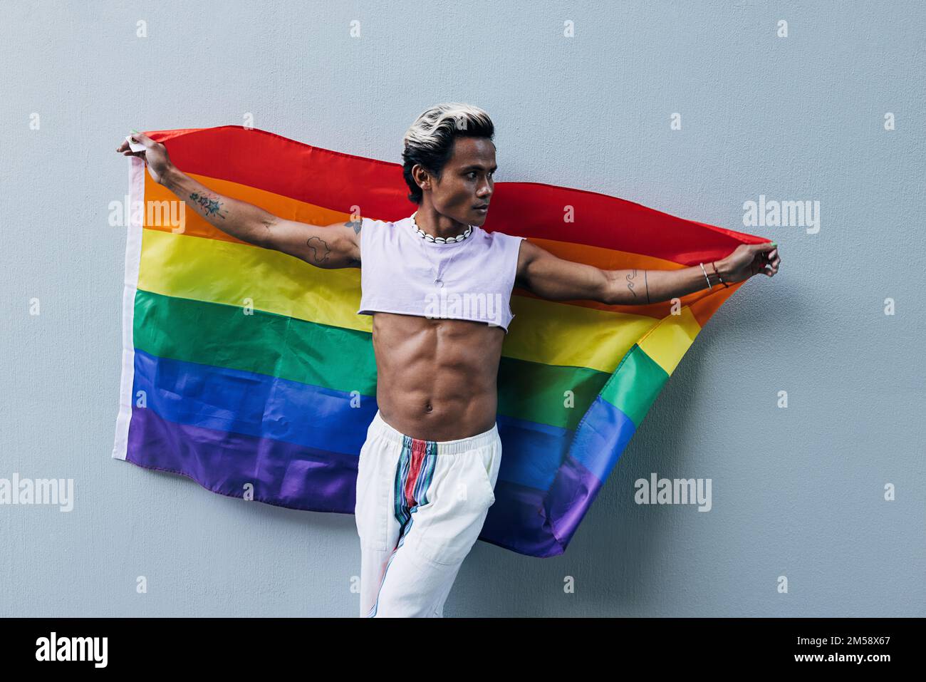 Un homme musclé et élégant portant un drapeau arc-en-ciel LGBT sur un mur gris Banque D'Images