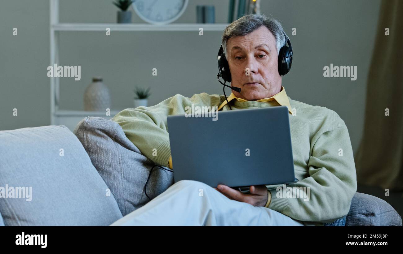 Senior 60s homme sur un canapé à la maison porter des écouteurs numériques parler en ligne à l'ordinateur portable web camera vieil homme utilisant l'application d'ordinateur pour la conférence discuter d'affaires Banque D'Images