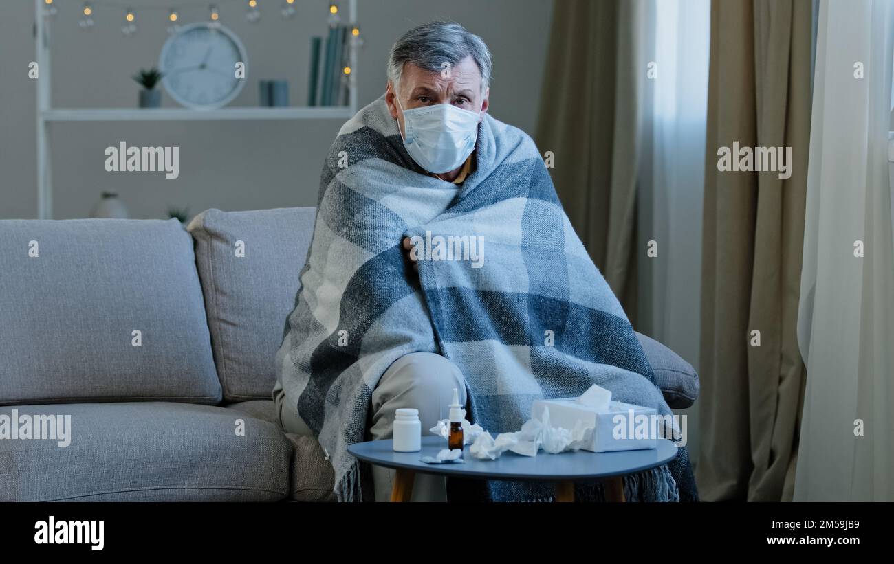 Malade vieux mâle dans le masque médical assis sur le canapé à la maison caucasien homme âgé enveloppé dans une couverture sentir mal maladie mature senior regardant Banque D'Images