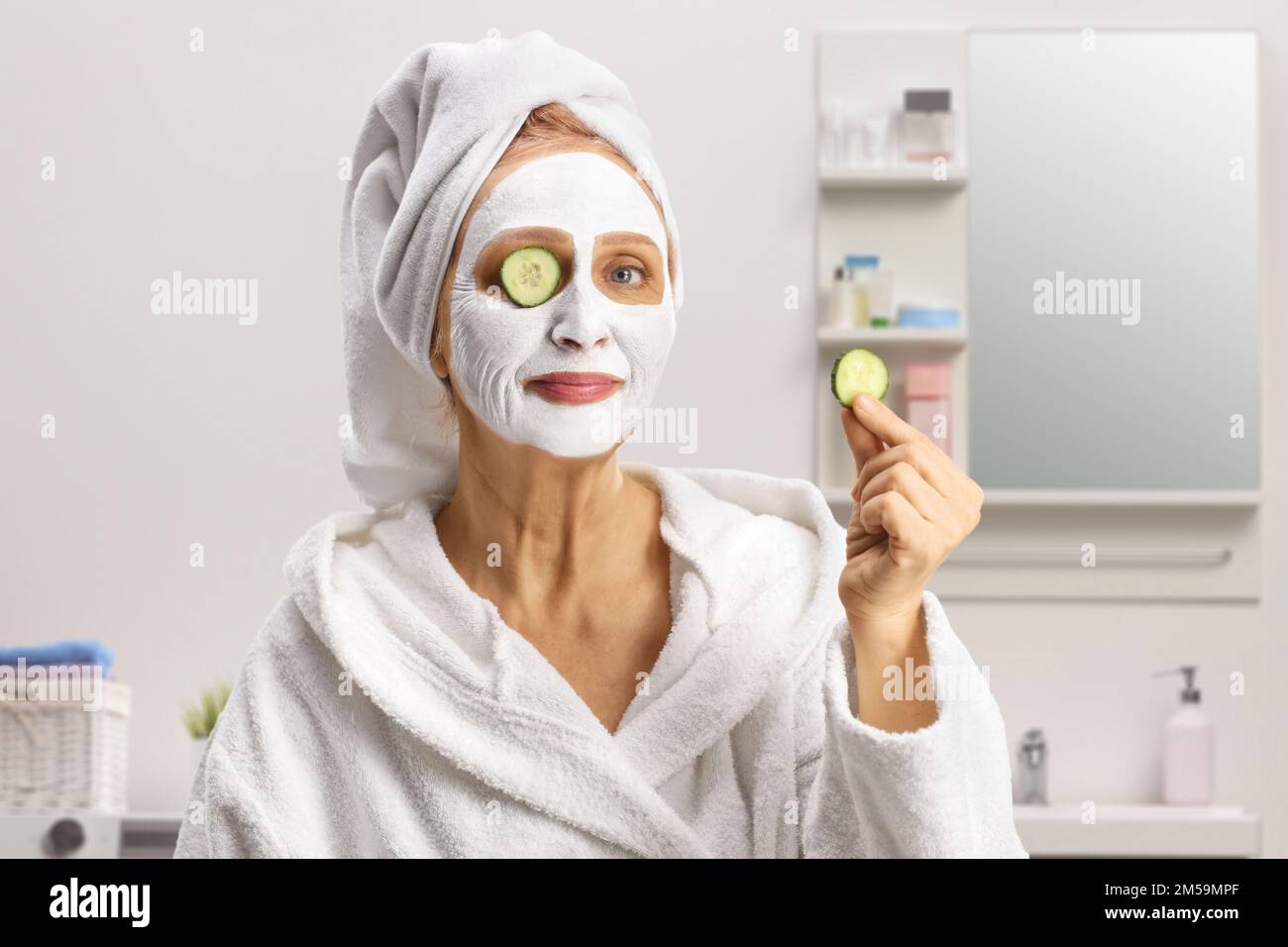 Femme avec cheveux dans une serviette et un masque de visage mettant le concombre sur les yeux à l'intérieur d'une salle de bains Banque D'Images
