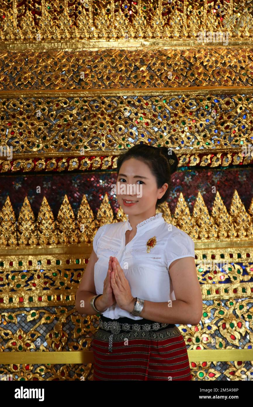 La dame thaïlandaise en costume thaïlandais met les paumes des mains ensemble en hommage dans le fond de mur de mosaïque de verre thaïlandais Banque D'Images
