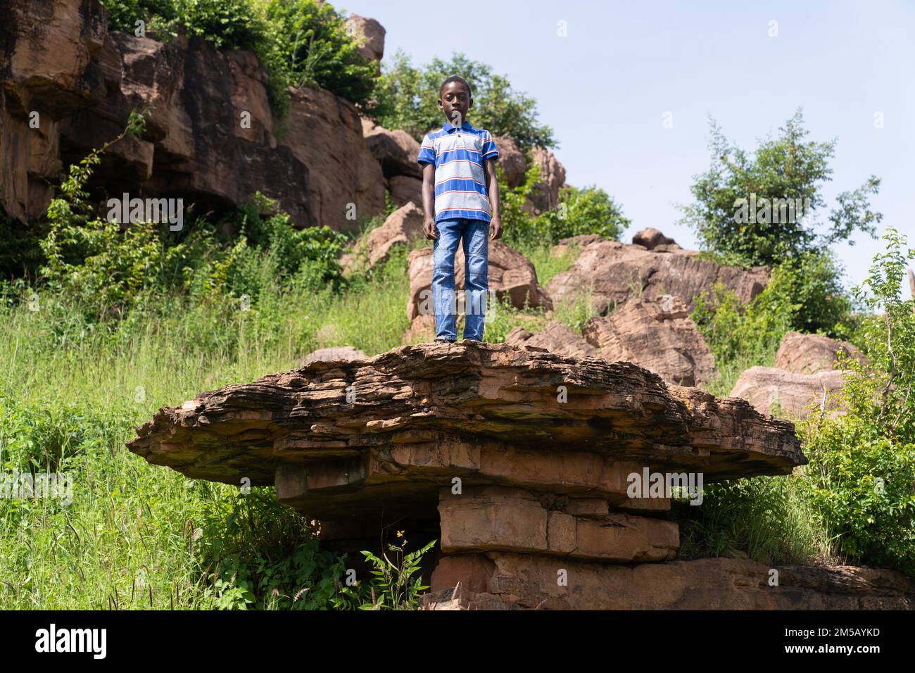 Mignon petit garçon de village africain dans un ensemble décontracté debout sur un rocher regardant l'appareil photo Banque D'Images