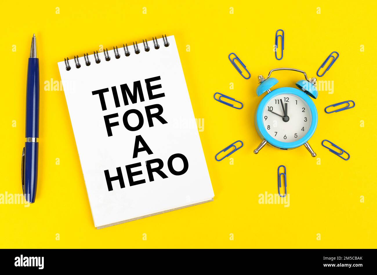 Concept d'entreprise. Sur une surface jaune il y a un réveil, un stylo et un bloc-notes avec l'inscription - temps pour un héros Banque D'Images Concept d'entreprise. Sur une surface jaune il y a un réveil, un stylo et un bloc-notes avec l'inscription - temps pour un héros Banque D'Images