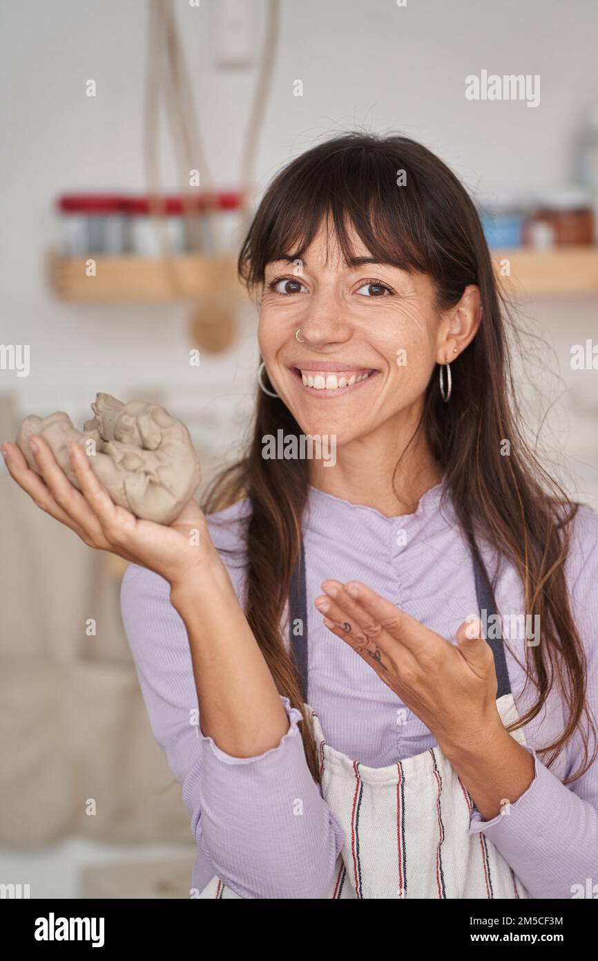 Femme en tablier regardant la caméra et souriant tout en posant avec de l'argile à la main dans le studio de poterie. Artisanat, art et passe-temps. Banque D'Images