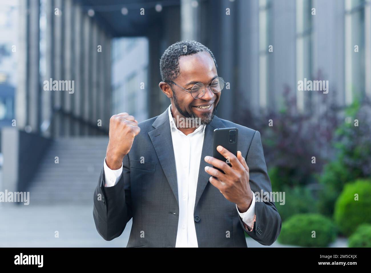 Homme d'affaires afro-américain patron à l'extérieur de l'immeuble de bureaux moderne utilisant le téléphone, homme senior célébrant la victoire succès lecture de bonnes nouvelles en ligne à partir de smartphone, tenant main vers le haut geste de triomphe . Banque D'Images