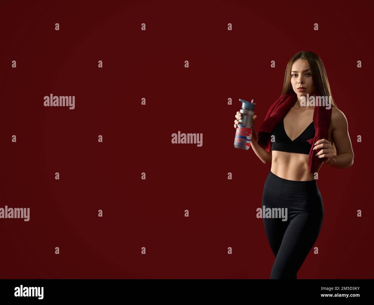 Vue avant de la femme entraîneur de fitness après l'entraînement avec une serviette autour du cou tenant l'agitateur avec de l'eau. Belle femme brune en noir posant avec la boisson aux protéines dans le studio rouge, regardant la caméra. Banque D'Images